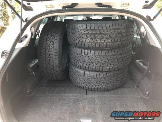 Four black tires stacked in the open trunk of a white SUV.