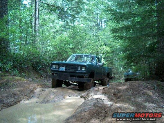 Green pickup truck driving through a muddy trail in the woods. Another vehicle is visible behind it