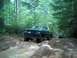 1979 Ford Bronco - A lifted green pickup truck navigates a muddy off-road path surrounded by trees, with another vehicle following.