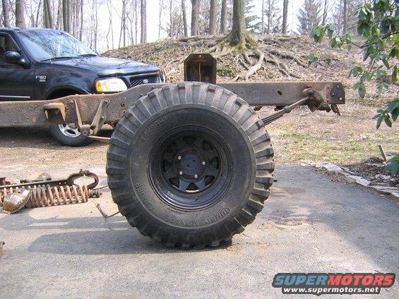Truck frame with large off-road tire, black pickup truck in background