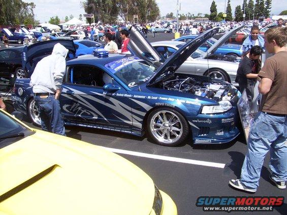Blue coupe with custom graphics and scissor doors, hood open showing engine. People looking at it. Yellow car in foreground