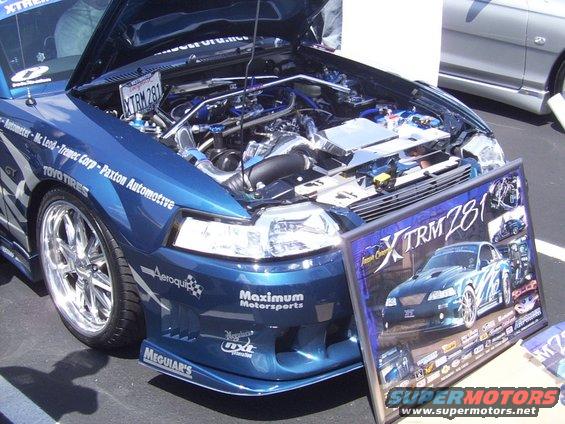 Blue Ford Mustang with hood open, chrome wheels, and custom engine modifications. Promotional poster for XTRM 281 is in the foreground