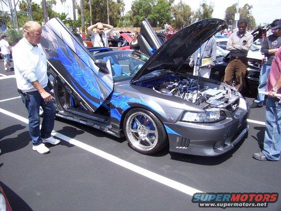 Gray Ford Mustang with blue flames and gullwing doors open, hood up showing engine. A man stands by the driver's door