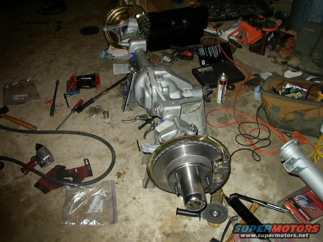 Vehicle rear axle assembly with brake rotors and calipers, surrounded by tools and parts on a garage floor