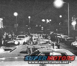 Cars on a street at night, some with headlights on. Watermark "SUPERMOTORS.NET" is visible