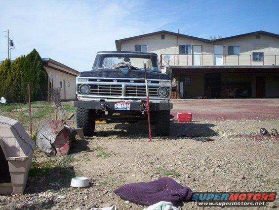 Black 1973 Ford F-Series truck parked in a gravel lot in front of a house. It has a red toolbox nearby