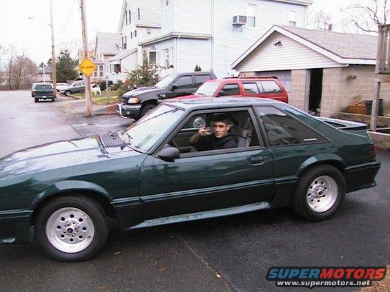 Green 1987 Ford Mustang coupe, driver giving peace sign, parked on asphalt with houses in background