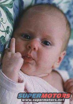 Baby with bald head and light skin, wearing a white shirt, gives the middle finger while lying on a patterned pillow