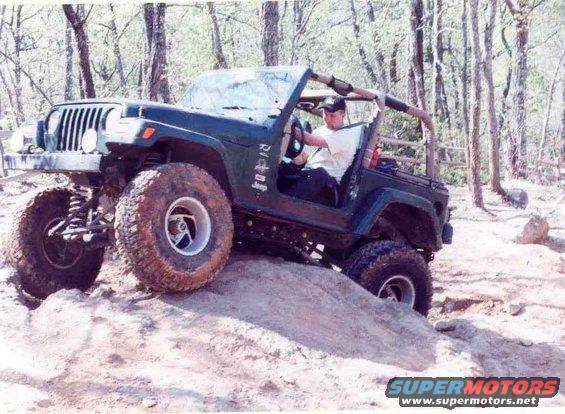 Green Jeep Wrangler climbing a dirt hill with large tires. Driver is visible
