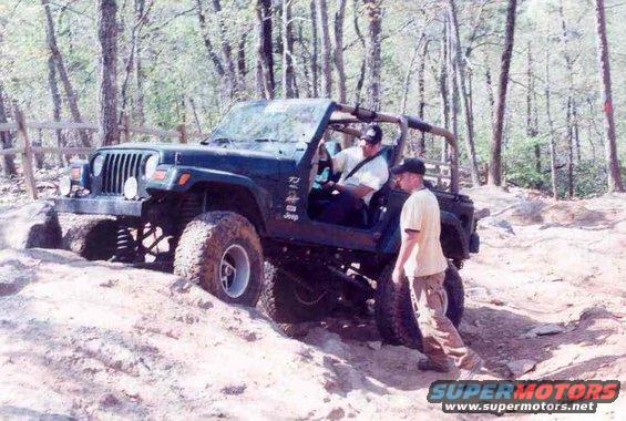 Dark green Jeep Wrangler off-roading on a dirt trail, with two men standing nearby