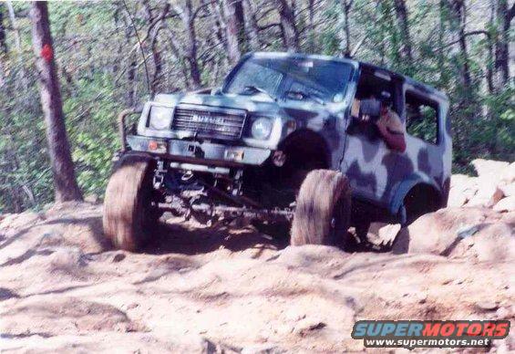 Camouflaged Suzuki Samurai off-roading on rocky terrain, front-left tire lifted