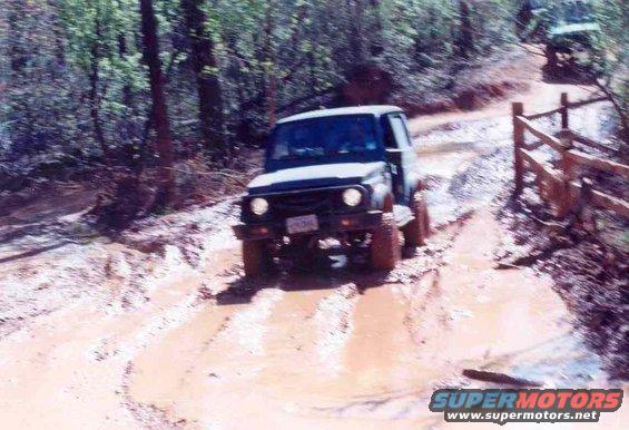 Suzuki Samurai driving through mud on a wooded trail. Another vehicle is visible further up the path