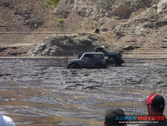 Two off-road vehicles are stuck deep in mud near a rocky hillside. People are watching from the foreground