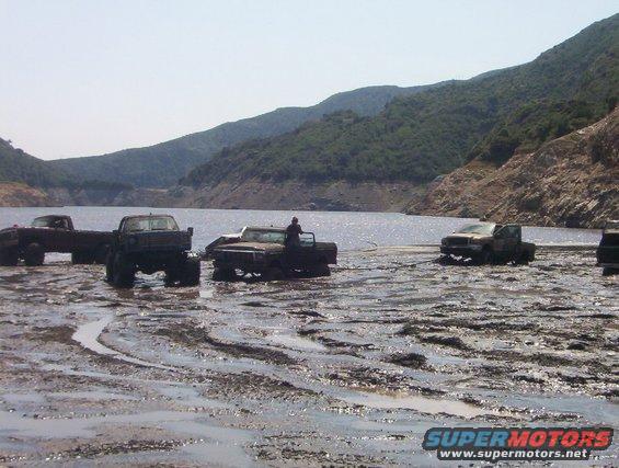 Several trucks are stuck in thick mud near a body of water. Hills are in the background