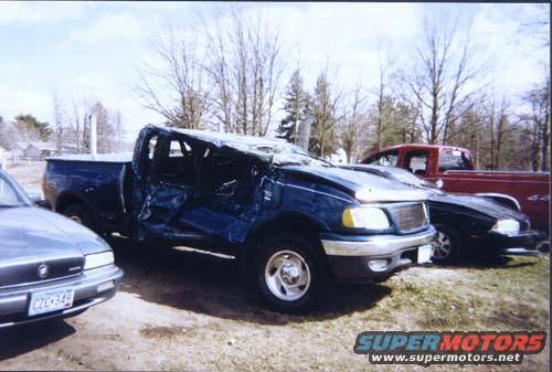 blue pickup truck with severe damage to the cab and roof, parked next to a red truck and a silver car