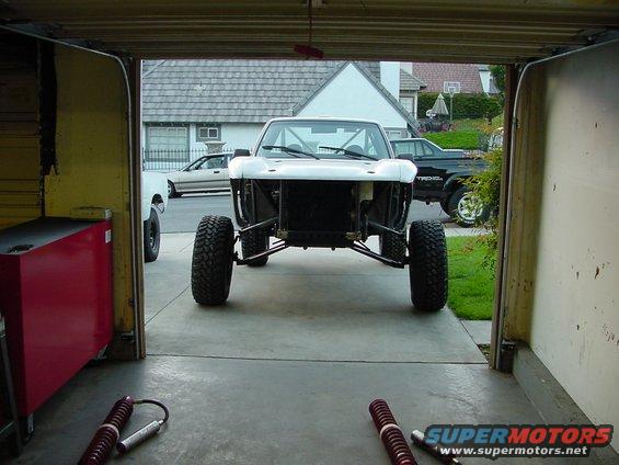 Front view of a custom off-road truck chassis with large tires, parked in a garage. Other vehicles are visible outside