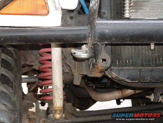 Front suspension with red coil spring, shock absorber, and part of a black bumper with a tow hook. Radiator visible behind bumper