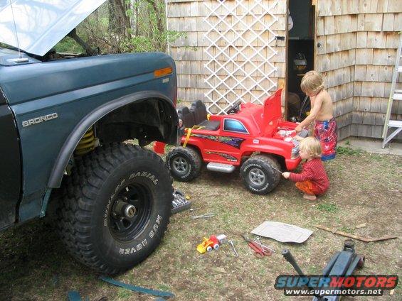 1986 Ford Bronco with hood up, large tires, next to a red toy truck where two children are playing
