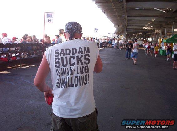 Man in white t-shirt with 'SADDAM SUCKS OSAMA bin LADEN SWALLOWS!' printed on back, standing in a crowd at an outdoor event