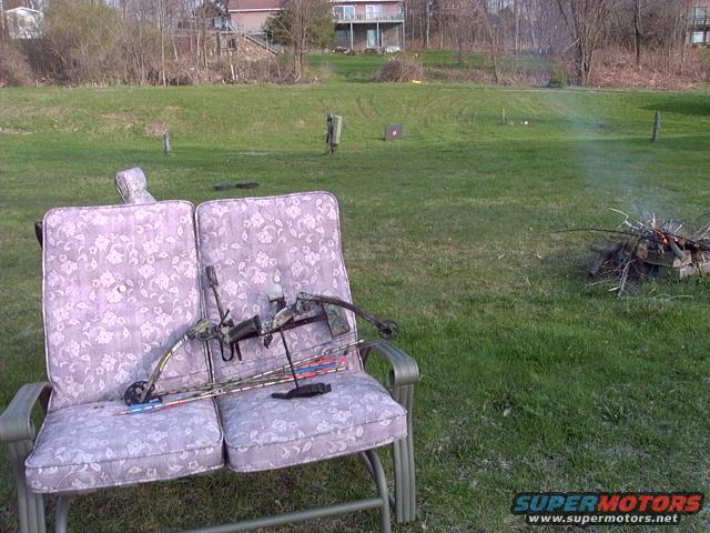 Compound bow and arrows resting on a floral-patterned chair in a grassy yard with a small campfire