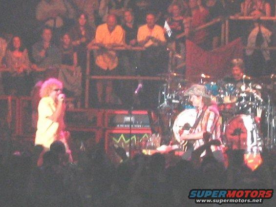Singer with curly hair on left, guitarist with hat on right, performing on stage with drums behind