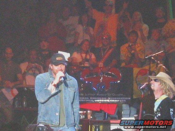 Stage with a singer in a denim jacket and a man in a cowboy hat, with a crowd in the background
