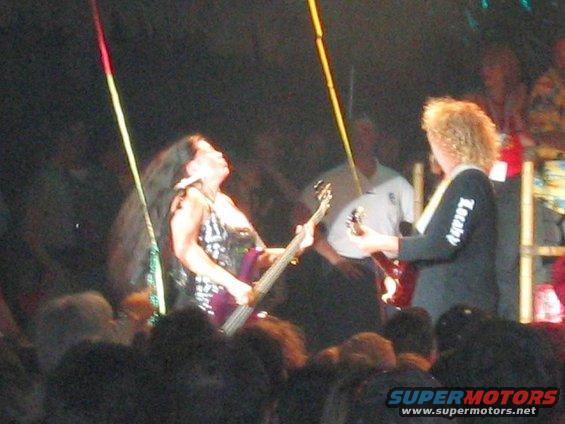 woman playing bass guitar on stage, another person plays guitar behind her, crowd in foreground