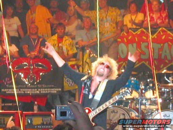 Sammy Hagar with blonde curly hair and sunglasses, wearing a black shirt, plays a red guitar on stage in front of a crowd and drums