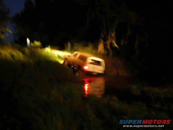 White SUV driving through water at night, rear view. Red taillights reflect in the water