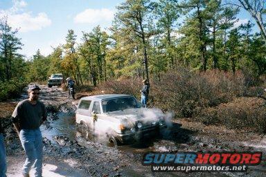 Truck stuck in a mud pit on a dirt road, trees all around. Another truck and a motorcycle are visible
