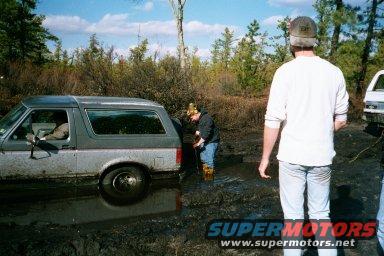 Gray Ford Bronco II stuck in mud with people around it. Another white vehicle is partially visible