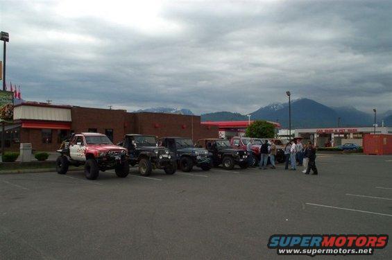 Several modified off-road trucks and SUVs parked in a lot near a building and mountains