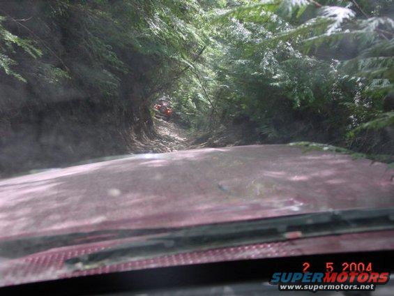 View from inside a vehicle driving on a muddy, wooded trail; another orange off-road vehicle is visible ahead