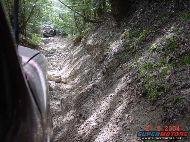 Two off-road vehicles on a rocky, muddy path in a forest, taken from inside a vehicle