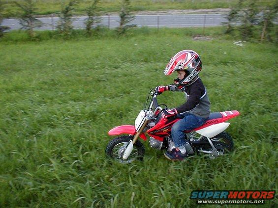 Child in helmet rides red dirt bike through grassy field
