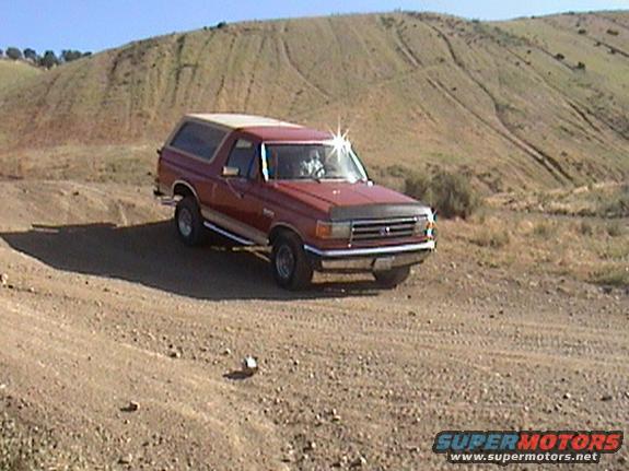 Red 1987 Ford Bronco with a tan top parked on a dirt road, facing right, with a hilly, dry landscape behind it
