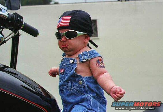 Baby in overalls and sunglasses sits on a motorcycle, wearing a bandana with an American flag patch