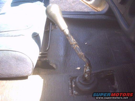 Gear shifter with worn rubber boot and taped handle on black ribbed floor mat inside a vehicle
