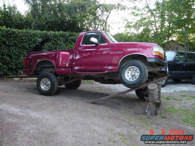 Red 1992 Ford F-150 pickup truck with a lifted suspension, angled on a wooden ramp and log