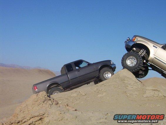 Gray pickup truck climbing a sand dune, with a lifted gold truck on a higher dune