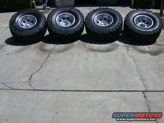 Four off-road tires with chrome rims lined up on a concrete driveway