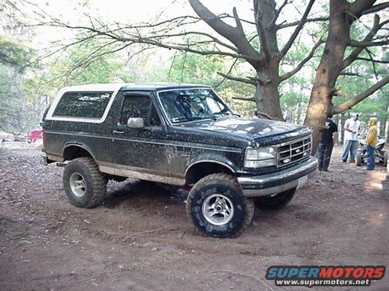 Dark green Ford Bronco with a white top, lifted suspension, and mud splatters, parked on a dirt trail