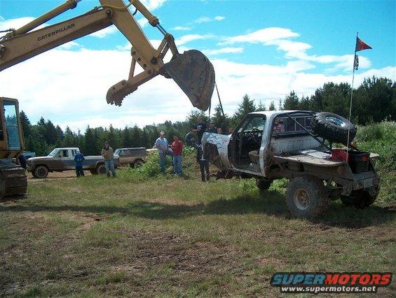 Excavator lifting a flatbed pickup truck. Other trucks and people are nearby in a grassy, wooded area