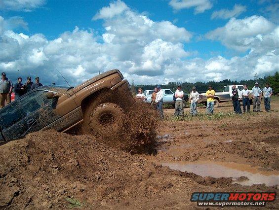Brown SUV stuck in mud, splashing dirt, with spectators watching