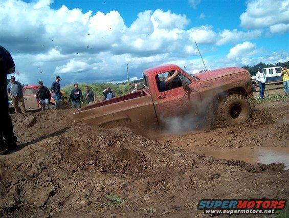 Red pickup truck stuck in mud pit with people watching. Another vehicle is visible in the background
