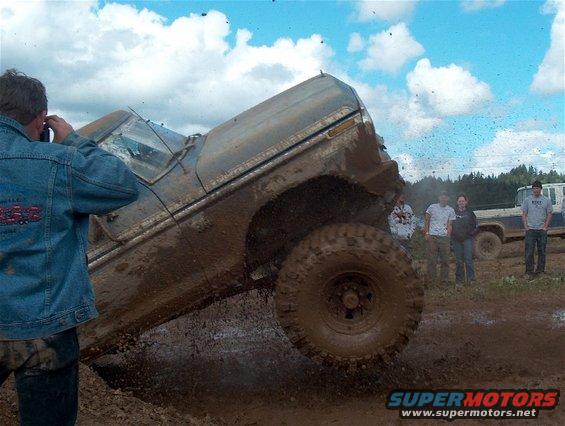 Muddy truck climbing out of a ditch with people watching. Another truck is parked in the background