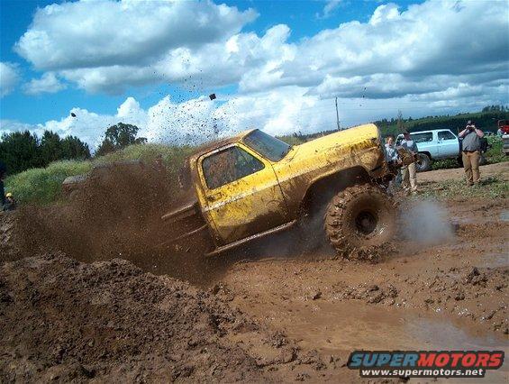 Yellow pickup truck with large tires driving through mud, splashing dirt. Another SUV is visible in the background