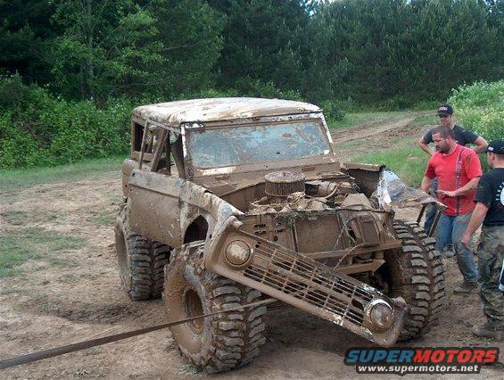 Muddy 1979 Ford Bronco with grille detached, hood open, and engine exposed. Three men stand nearby