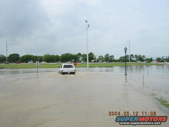 White pickup truck driving through floodwaters on a road, with other vehicles visible in the distance