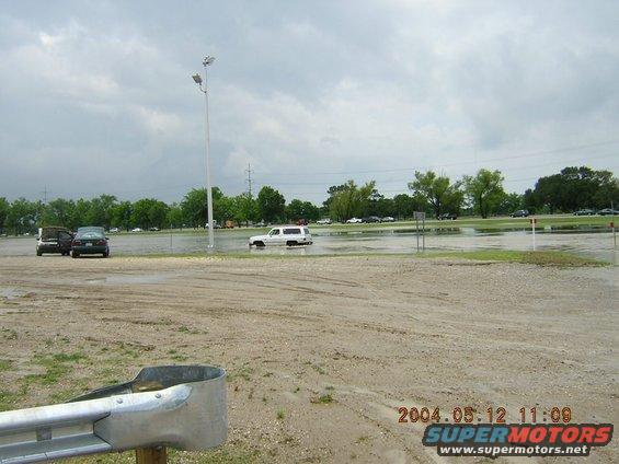 White SUV partially submerged in floodwater, seen from a muddy foreground with a metal guardrail